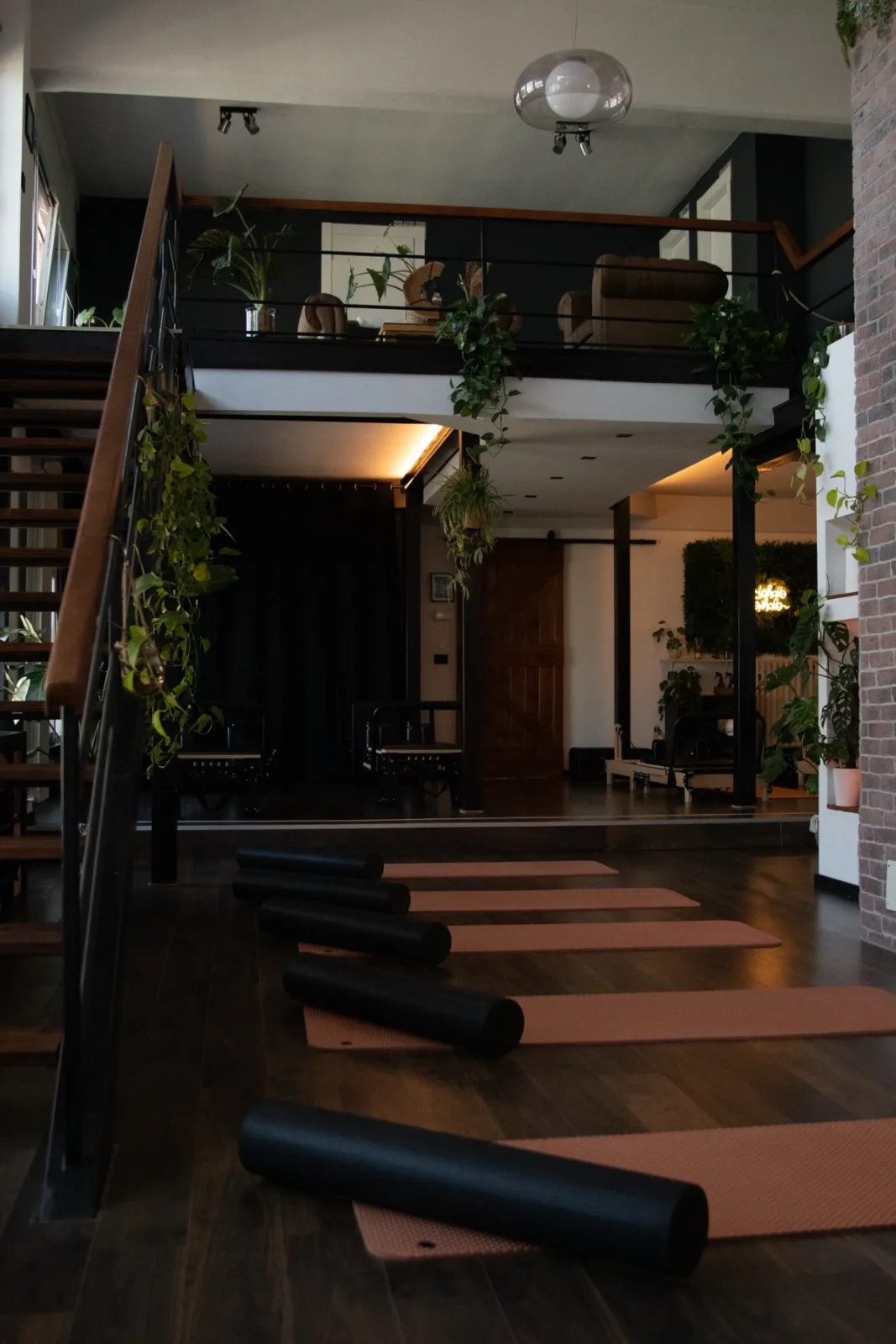 Vista d'insieme dello studio su doppio livello: il soppalco con piante e poltrona sopra, i tappetini terracotta e i reformer allineati sotto, l'insegna al neon IL LOFT sullo sfondo.