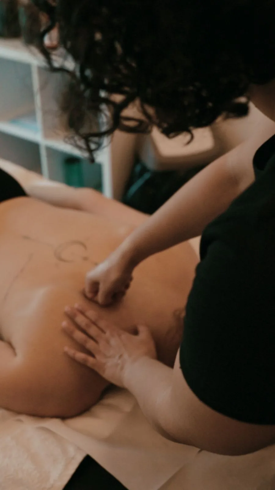 La massoterapista lavora con entrambe le mani sulla schiena della cliente, tra scaffali e boccette dello studio sullo sfondo.